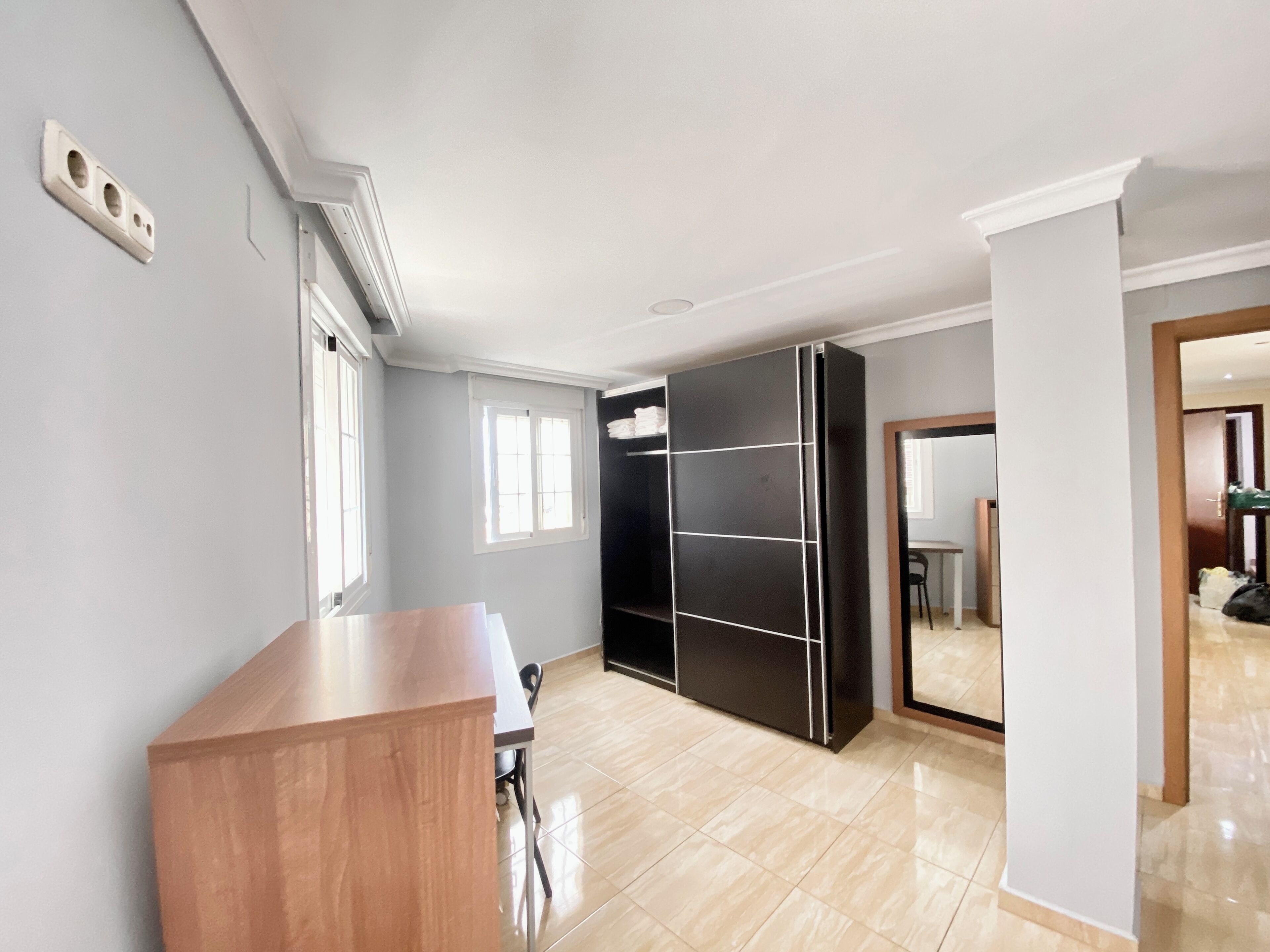 Una habitación con un escritorio de madera, un armario negro, una ventana y una puerta que da a otra estancia.