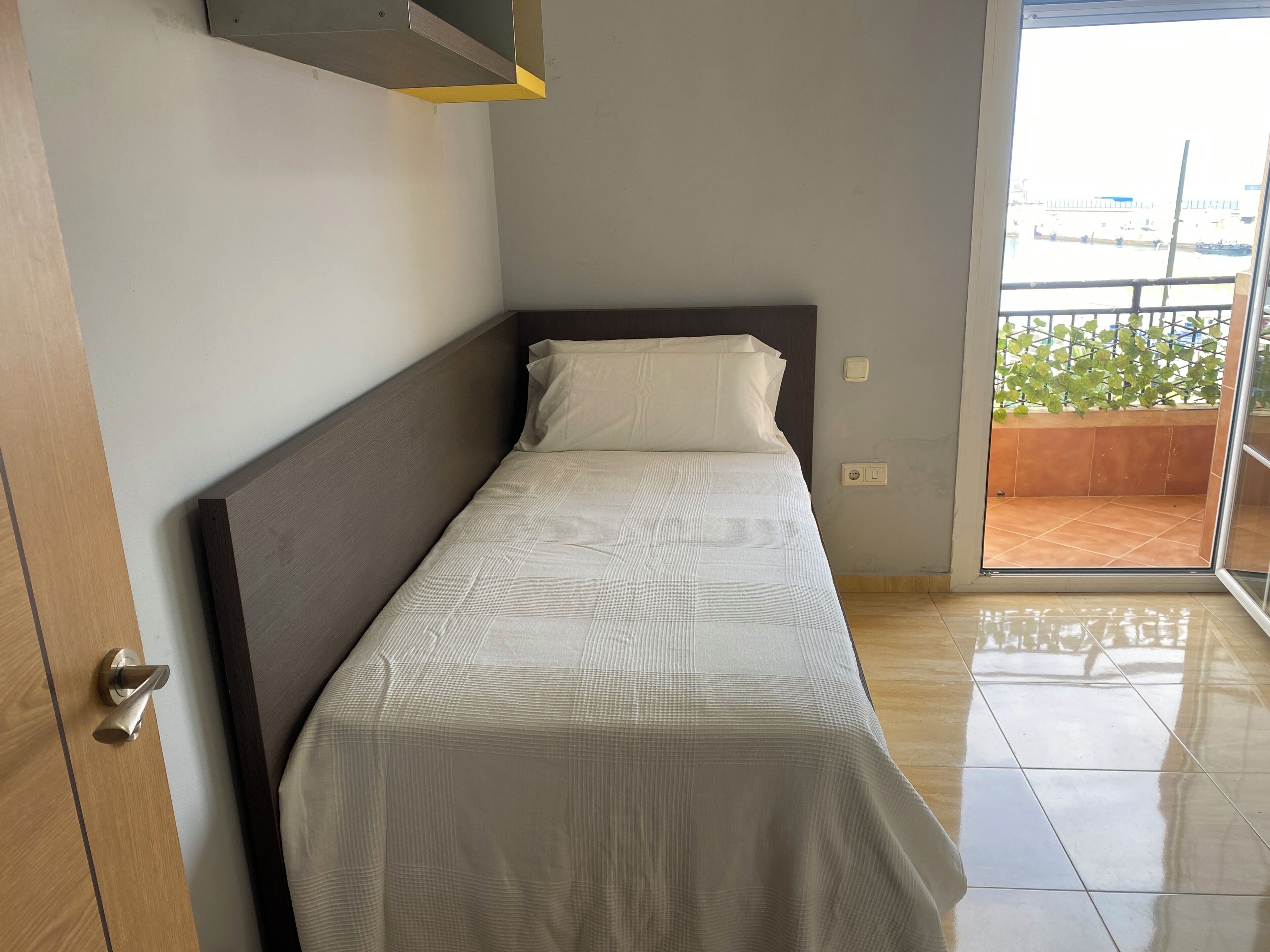 Una cama individual con cabecera de madera en una habitación con piso de baldosas y vista a un balcón con vegetación.