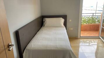 Una cama individual con cabecera de madera en una habitación con piso de baldosas y vista a un balcón con vegetación.