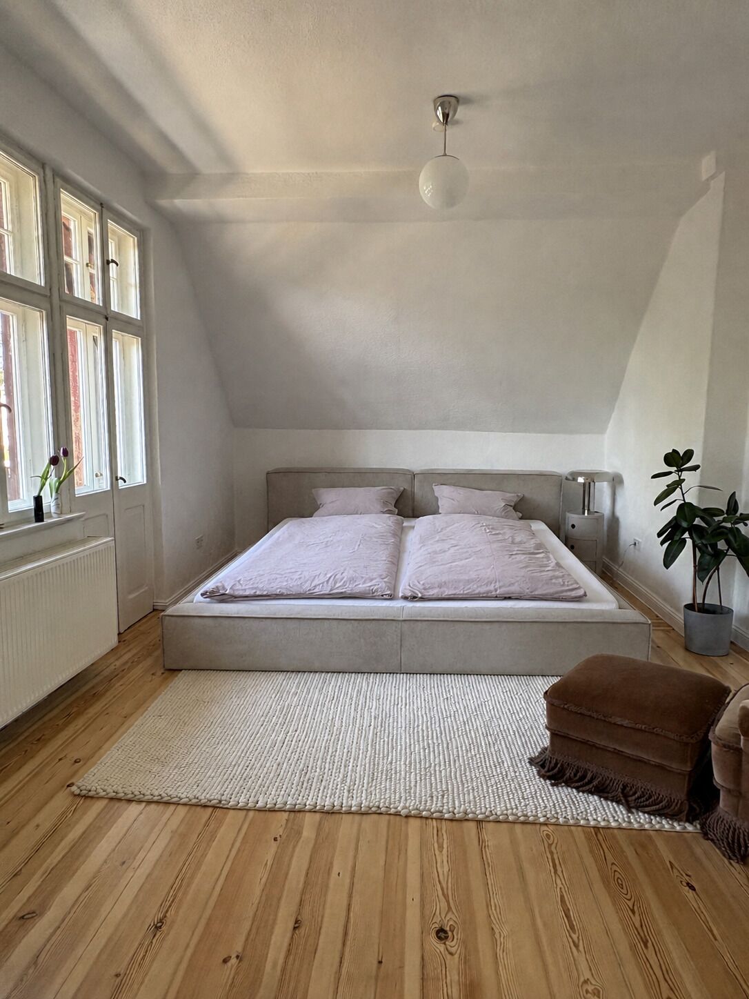 A bedroom with a large bed, a bedside table, a nightstand, a window with curtains, and a potted plant.