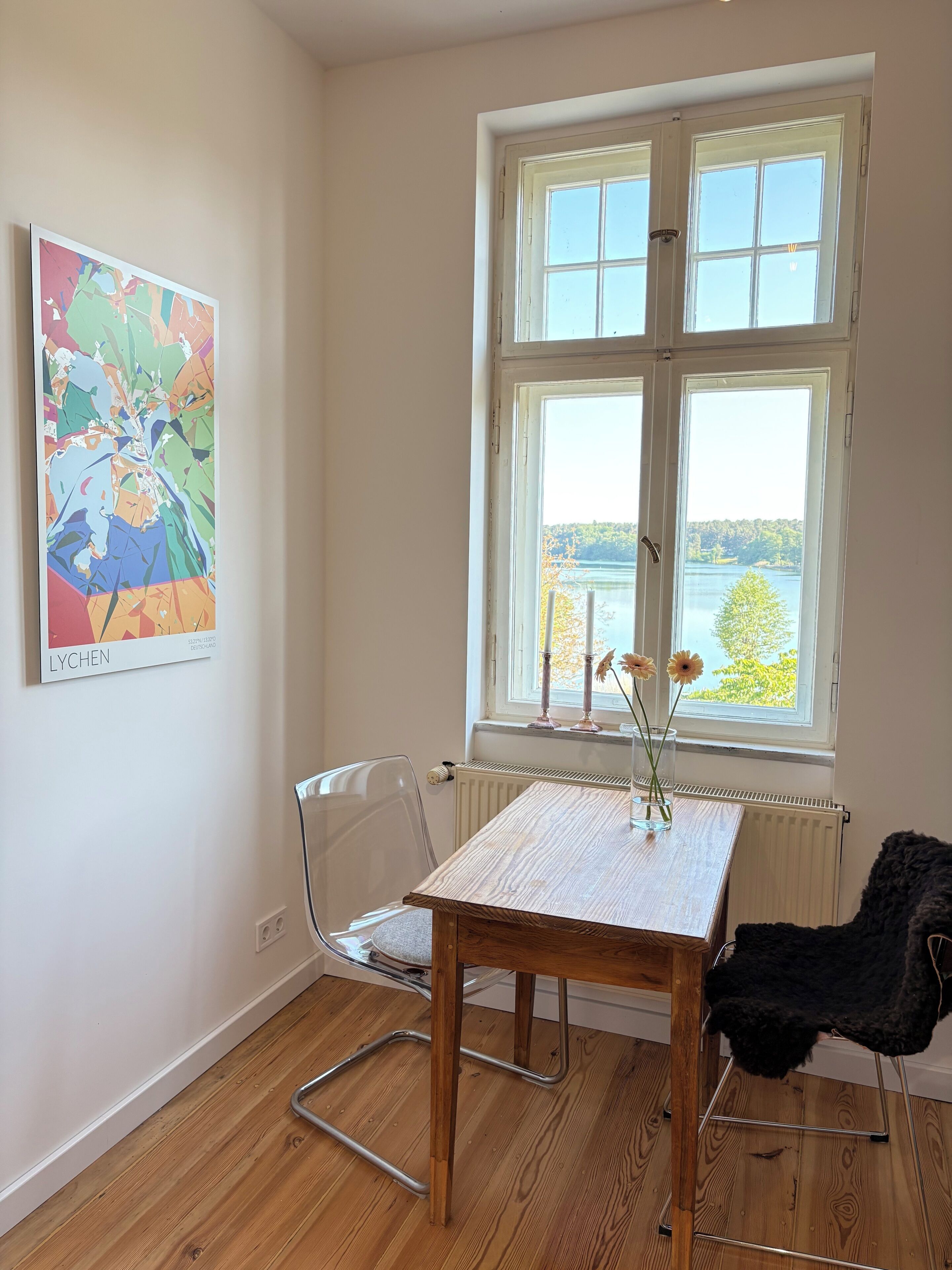 A room with a wooden table, a chair, a radiator, and a window with a view of a lake and trees.