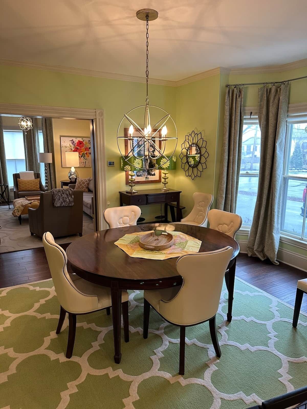 A dining area with a round table, six chairs, a chandelier, and a view of the outdoors through a window.