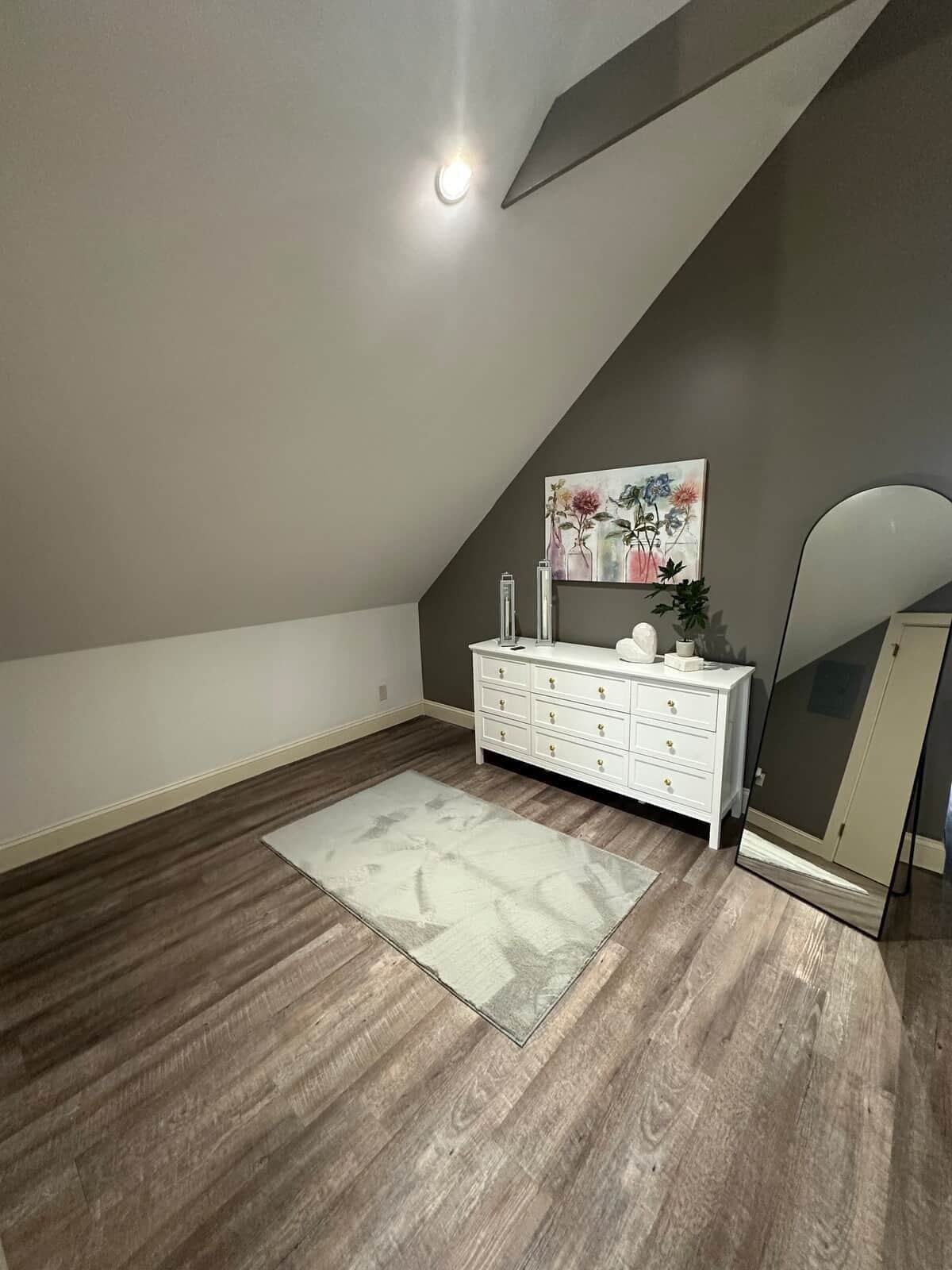 A room with a sloped ceiling, a white dresser with drawers, a painting on the wall, and a large mirror.