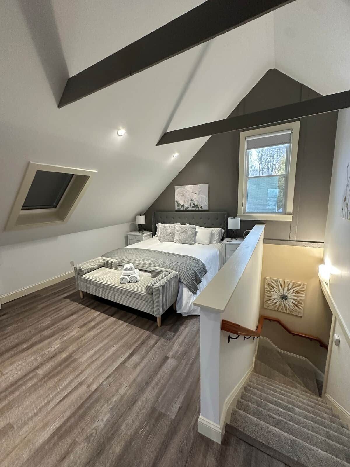 Ein Schlafzimmer mit schrägem Dach, einem Bett mit Kopfteil, einem Fenster und einer Treppe.