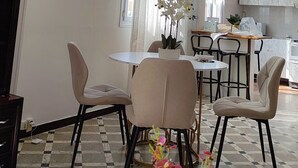 A dining area with a round table, chairs, and a vase with flowers. There is a kitchen in the background with cabinets and a countertop.