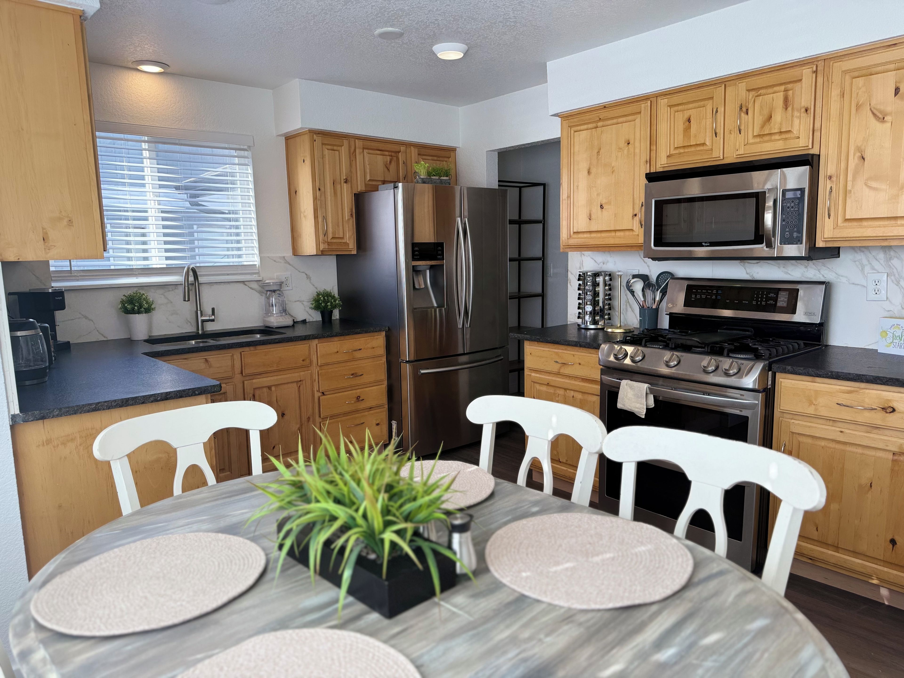 A modern kitchen with wooden cabinets, stainless steel appliances, a dining table with chairs, and a window with blinds.