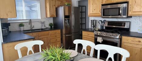 A modern kitchen with wooden cabinets, stainless steel appliances, a dining table with chairs, and a window with blinds.