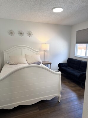 A bedroom with a white bed, a bedside table, a lamp, a dark-colored sofa, and a window with blinds. - Modern Home/ Near DIA & Anschutz Medical Campus (Denver)