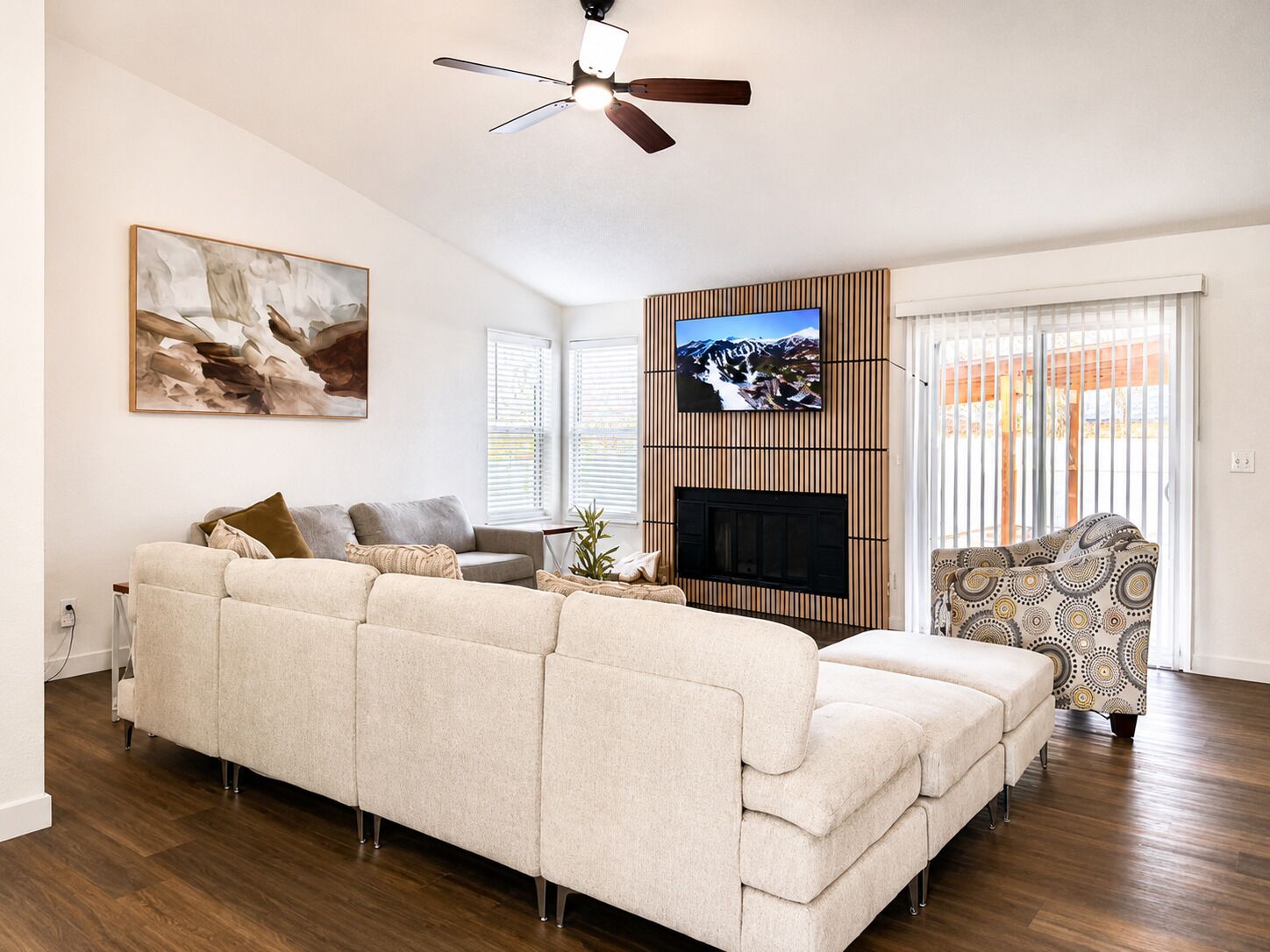 A living room with a sectional sofa, a flat-screen TV mounted on a wooden wall, a fireplace, and a ceiling fan.
