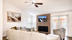 Ein Wohnzimmer mit Ecksofa, Flachbildfernseher an einer Holzwand montiert, Kamin und Deckenventilator.