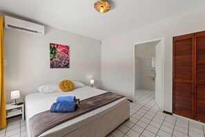 A hotel room with a bed, bedside lamps, a wall-mounted air conditioner, a picture of a cactus, and a wooden door. - Villa Cuartillo | Spacious 8-person villa with a private pool (Willemstad)
