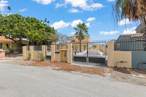 A gated residential area with a paved driveway, a palm tree, and houses with tiled roofs. - Villa Cuartillo | Spacious 8-person villa with a private pool (Willemstad)