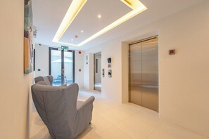 A modern lobby with a reception desk, a waiting area with chairs, an elevator, and a glass door leading outside. - Bellevue Beachfront Apartments (Choeng Thale)