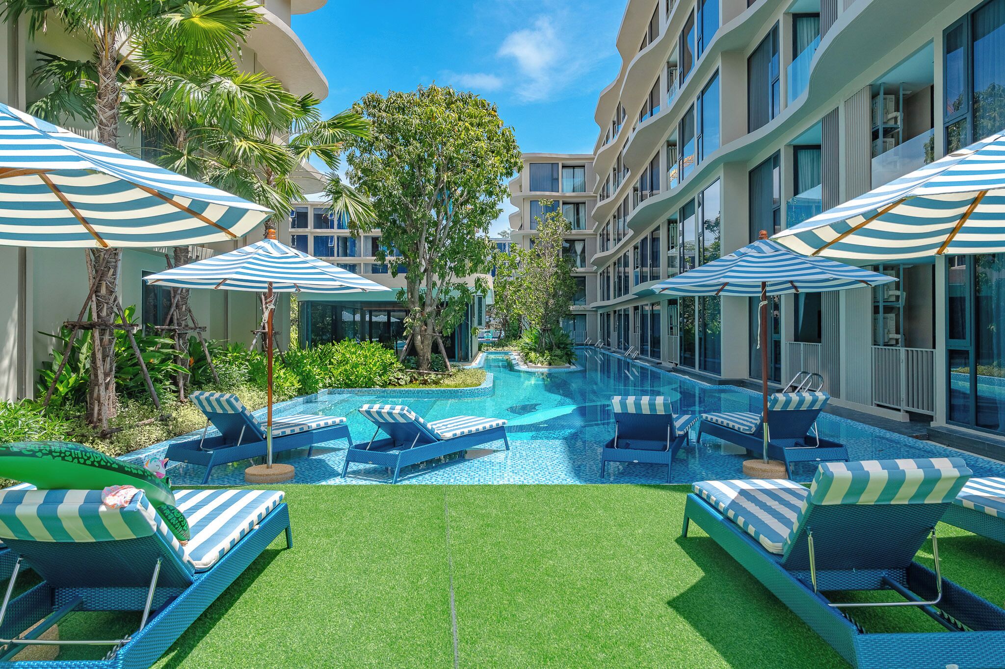 Ein Hotelpoolbereich mit mehreren Liegestühlen und Sonnenschirmen, umgeben von Grünflächen und moderner Architektur.
