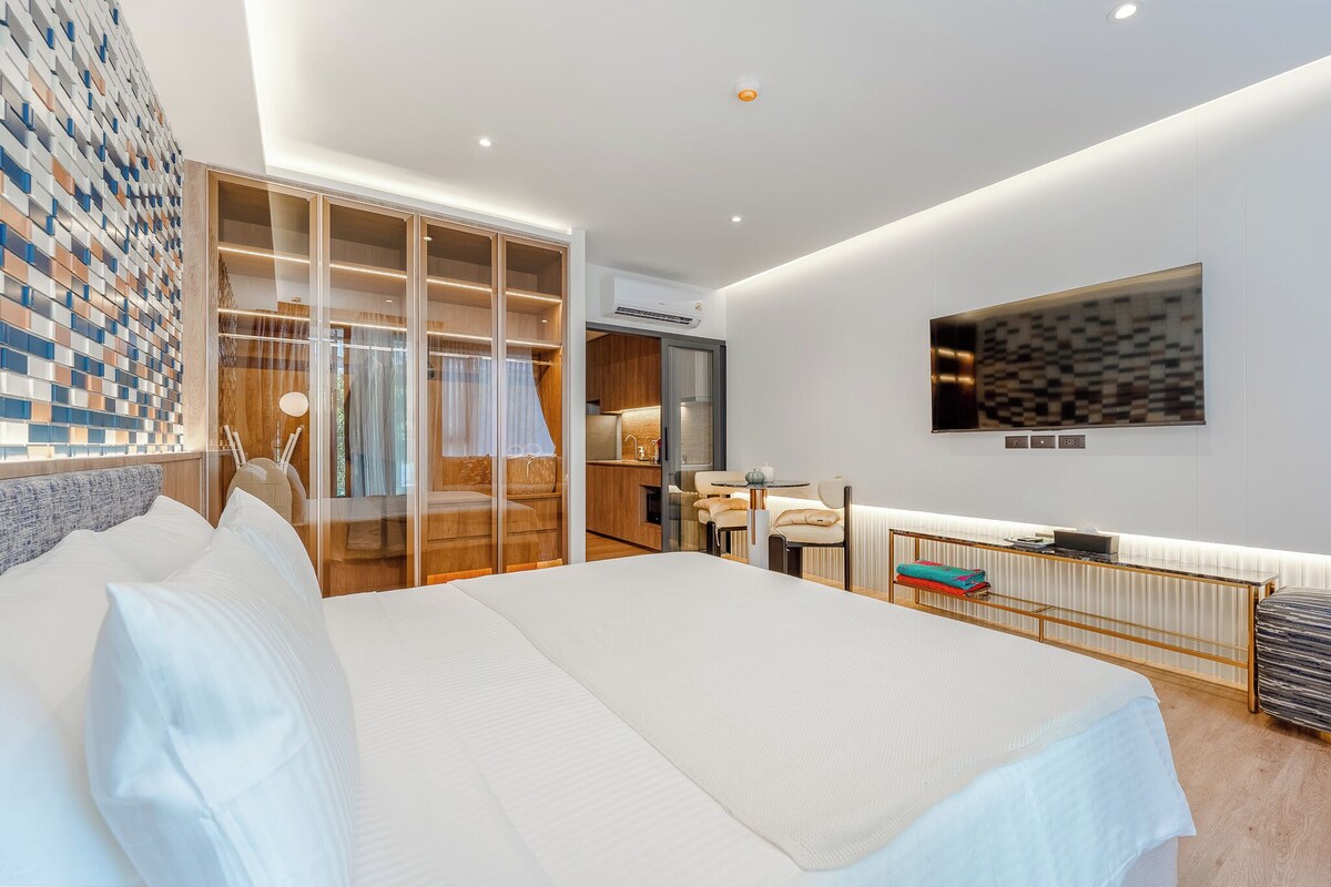 A modern hotel room with a large bed, a walk-in closet, a kitchenette, and a flat-screen TV mounted on the wall.
