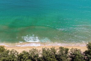 An aerial view of a beach with clear turquoise water, gentle waves, and dense green foliage in the foreground. - Bellevue Beachfront Apartments (Choeng Thale)