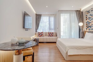 A modern hotel room with a bed, sofa, dining table, and a large window with curtains. - Bellevue Beachfront Apartments (Choeng Thale)