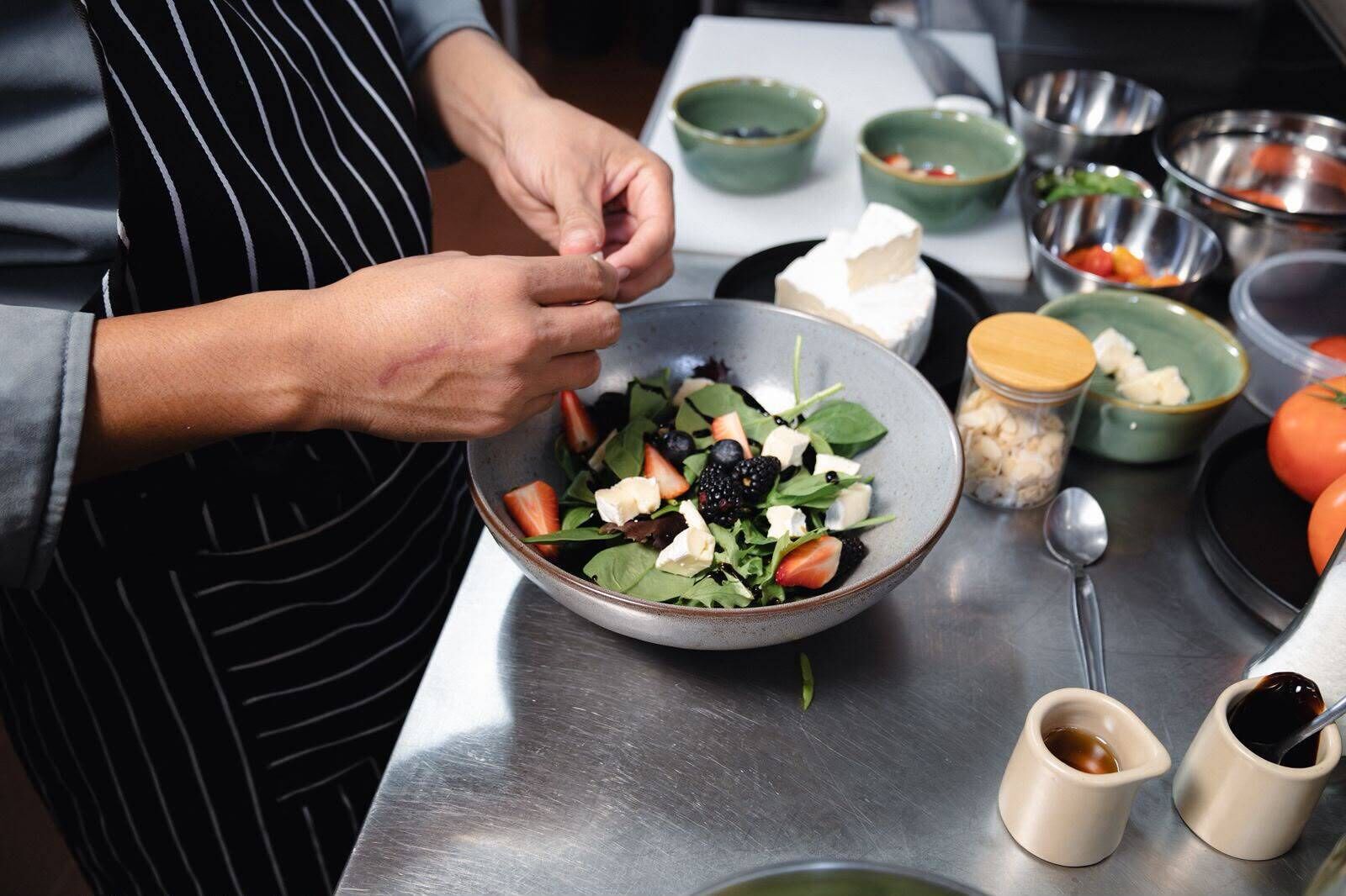 A person preparing a salad with various ingredients and kitchen utensils on a stainless steel countertop.