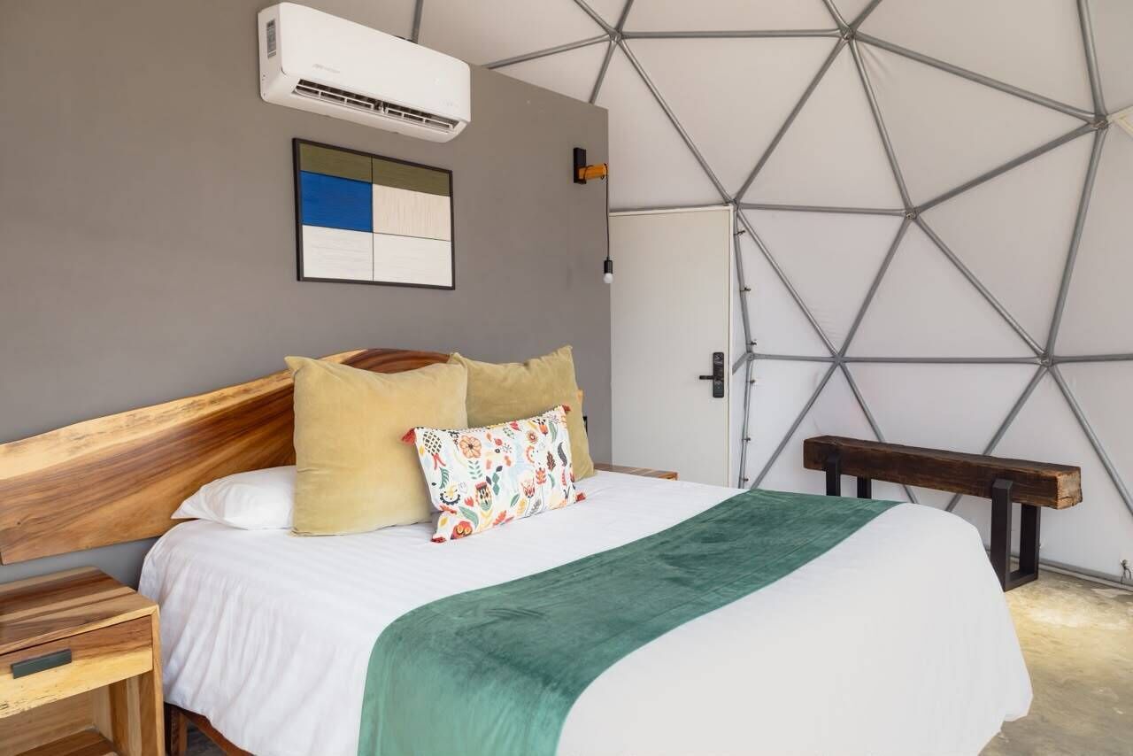 A bedroom with a wooden headboard, a bed with white bedding and a green throw, a bench, and a geometric wall design.