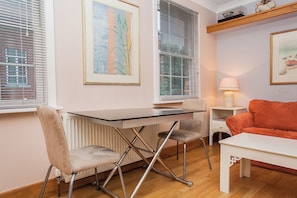 A room with a glass-top table, two chairs, a red sofa, a white side table with a lamp, and two framed artworks on the wall. - Sunlit 1BDR Near Westminster and Big Ben (London)