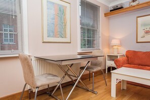 Ein Zimmer mit einem Glastisch, zwei Stühlen, einem roten Sofa, einem weißen Nachttisch mit Lampe sowie zwei gerahmten Kunstwerken an der Wand.