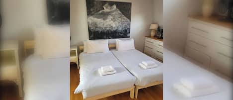 A hotel room with two beds, a wooden floor, a painting on the wall, a bedside table with a lamp, and a dresser.