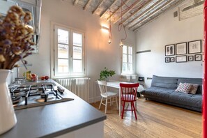 Eine moderne Küche mit Gasherd, ein Essbereich mit weißem Tisch und Stühlen, ein graues Sofa und ein roter Sessel.