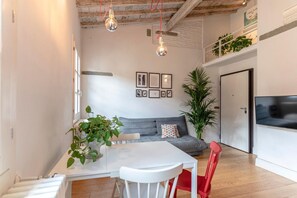 Een moderne woonkamer met een grijze bank, een witte eettafel en een rode fauteuil. Er hangen twee hanglampen, staat een grote potplant en is een flatscreen televisie aan de muur bevestigd.