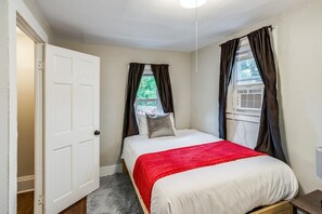 Un dormitorio con cama, una puerta blanca, una ventana con cortinas y un equipo de aire acondicionado.