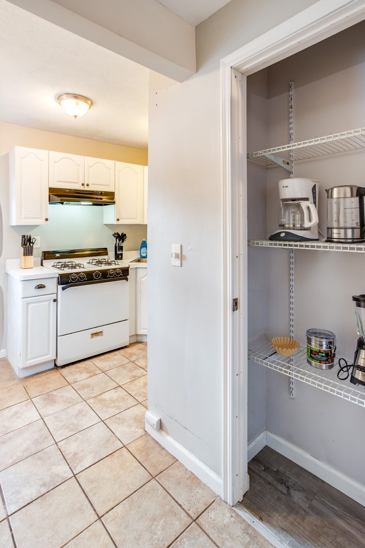 A kitchen with white cabinets, a stove, and a coffee maker on a shelf.