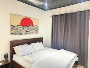 A neatly made bed with white linens and a rolled towel, a wooden headboard, a wall-mounted artwork depicting a red sun and waves, and a grey curtain. - City Centre Oasis (Accra)