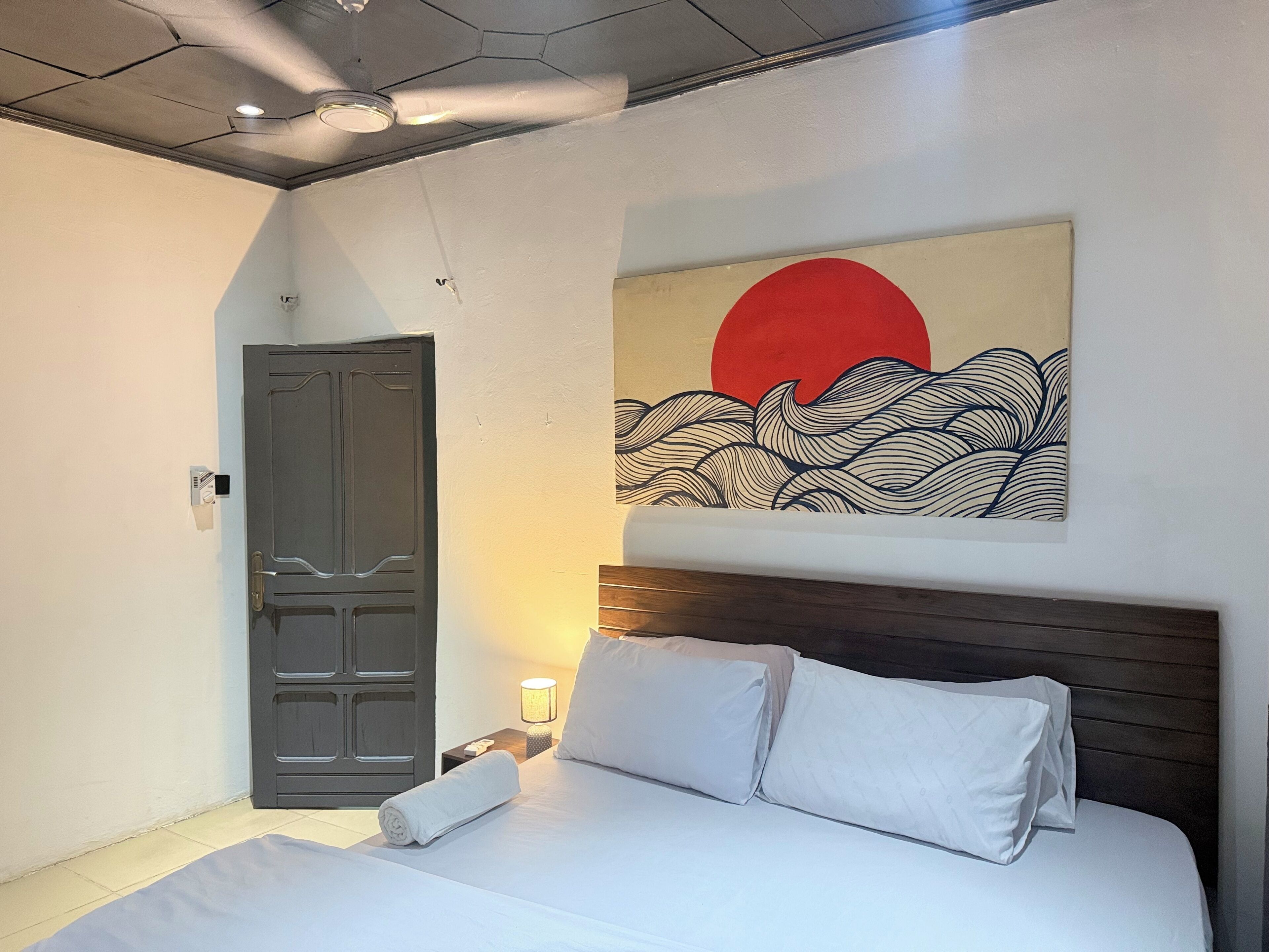 A bedroom with a bed, a door, a wall-mounted artwork, and a nightstand with a lamp.