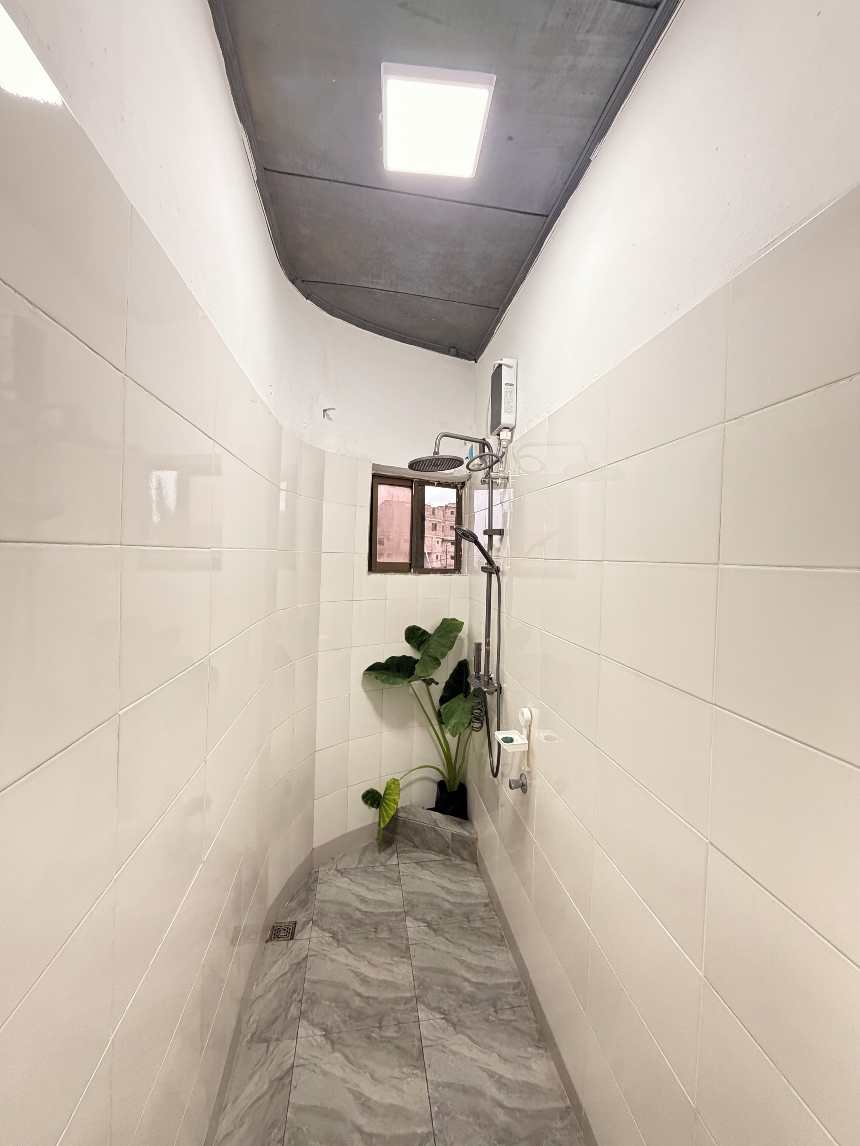 A narrow shower area with white tiled walls, a marble floor, a ceiling light, a handheld showerhead, a potted plant, and a small window.