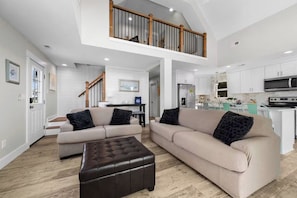 A spacious living area with a high ceiling, a staircase, and a kitchen area. - The Salted Rim | Sound Sunsets • 2-Min Walk to Beach • Bikes • Family Fun (Surf City)