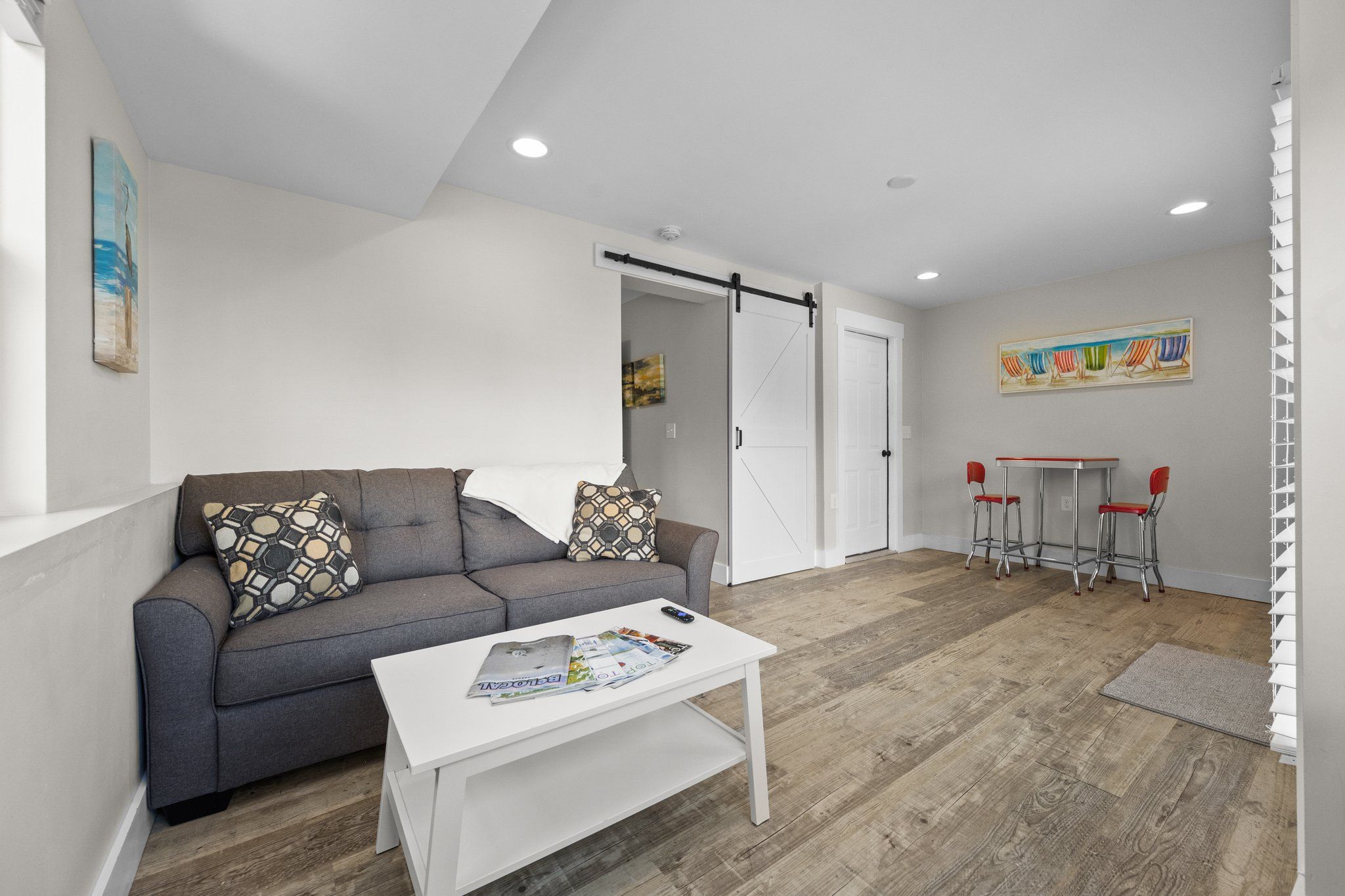 A modern living room with a gray sofa, a white coffee table, and a sliding door leading to a dining area with red chairs and a small table.