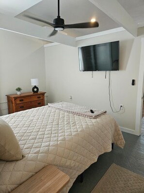 A bedroom with a bed, a ceiling fan, a TV mounted on the wall, a nightstand, and a bedside table.