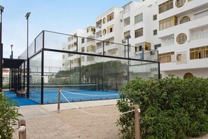 Ein Tennisplatz, umgeben von einer Glasumrandung und einem Gebäude mit Balkonen.