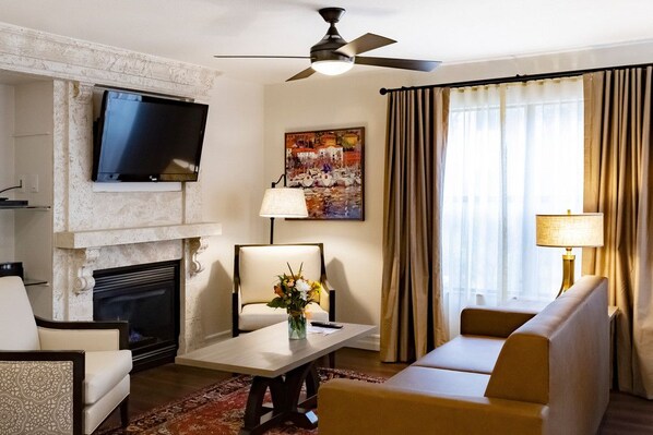 A living room with a fireplace, a flat-screen TV mounted above it, a sofa, a coffee table with a vase of flowers, and a ceiling fan.