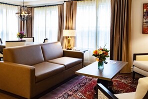 A hotel room with a sofa, a small table with a vase of flowers, a lamp, and a view of the outdoors through large windows with curtains.