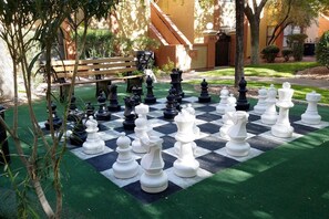 A large outdoor chessboard set up on artificial grass with surrounding greenery and a bench.