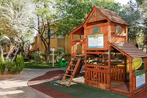 A wooden playhouse with a slide and a ladder, surrounded by greenery and a paved area.