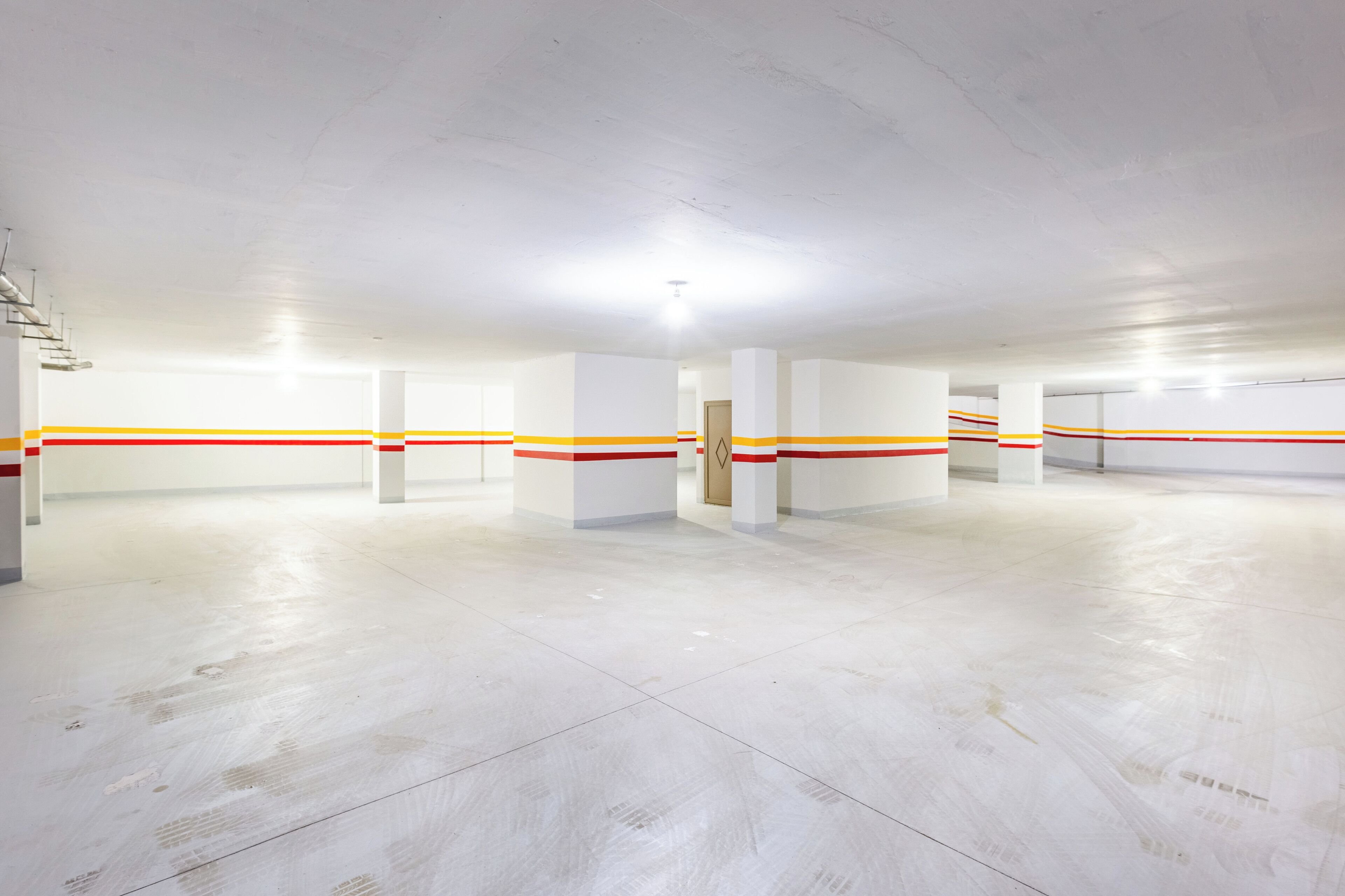 Un vaste espace de stationnement intérieur aux murs blancs et au sol en béton, agrémenté de nombreuses colonnes et d’une porte.