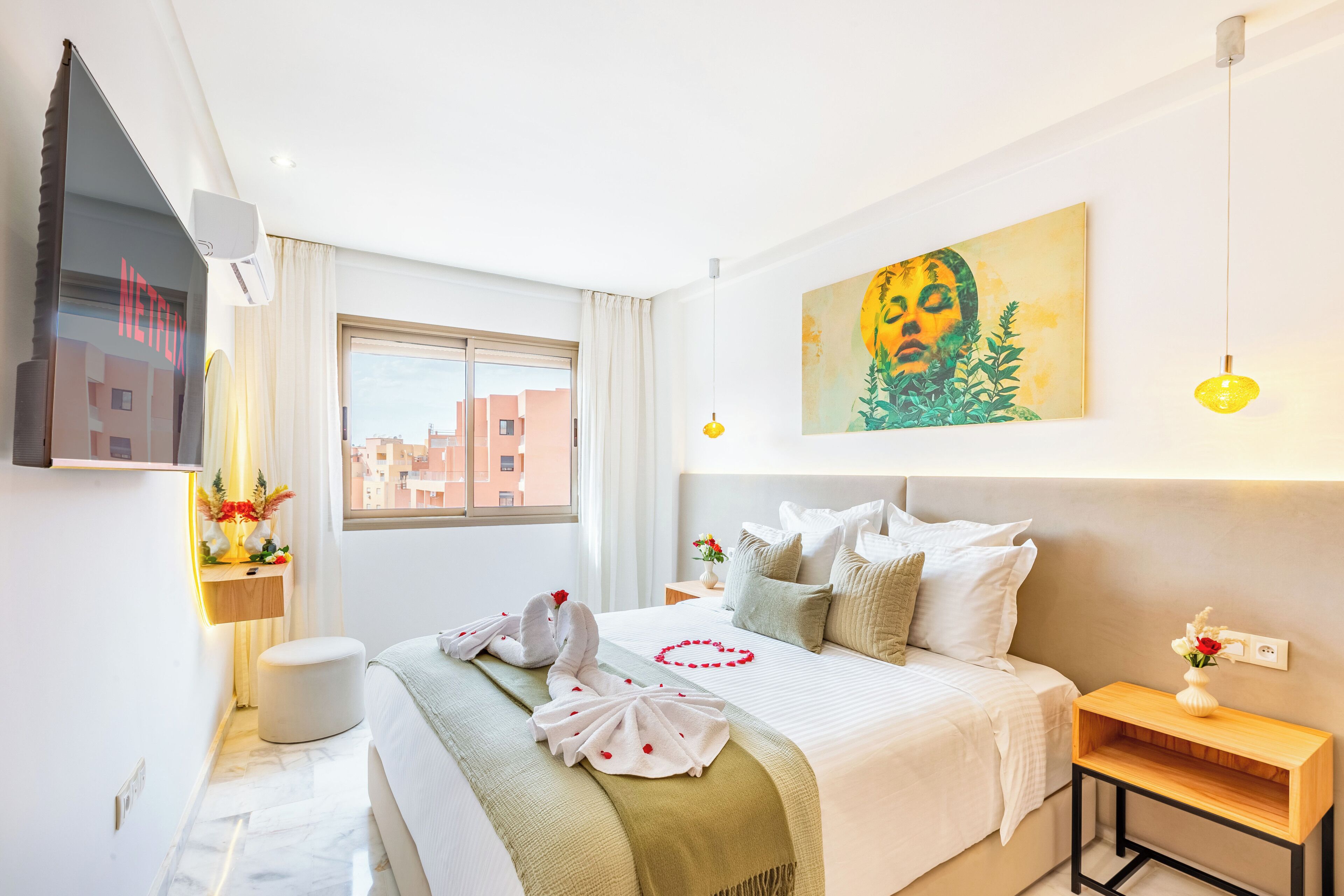 Una habitación de hotel con cama, mesita de noche, televisor montado en la pared, ventana y un cuadro.