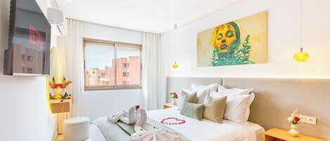 Una habitación de hotel con cama, mesita de noche, televisor montado en la pared, ventana y un cuadro.