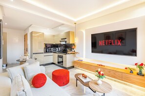 A modern living room with a sofa, ottoman, and a coffee table. A kitchen is visible in the background with a microwave and oven. - Cosy 1BR Apartment in Guéliz, Free Parking, Near Carré Eden Mall (Marrakesh)
