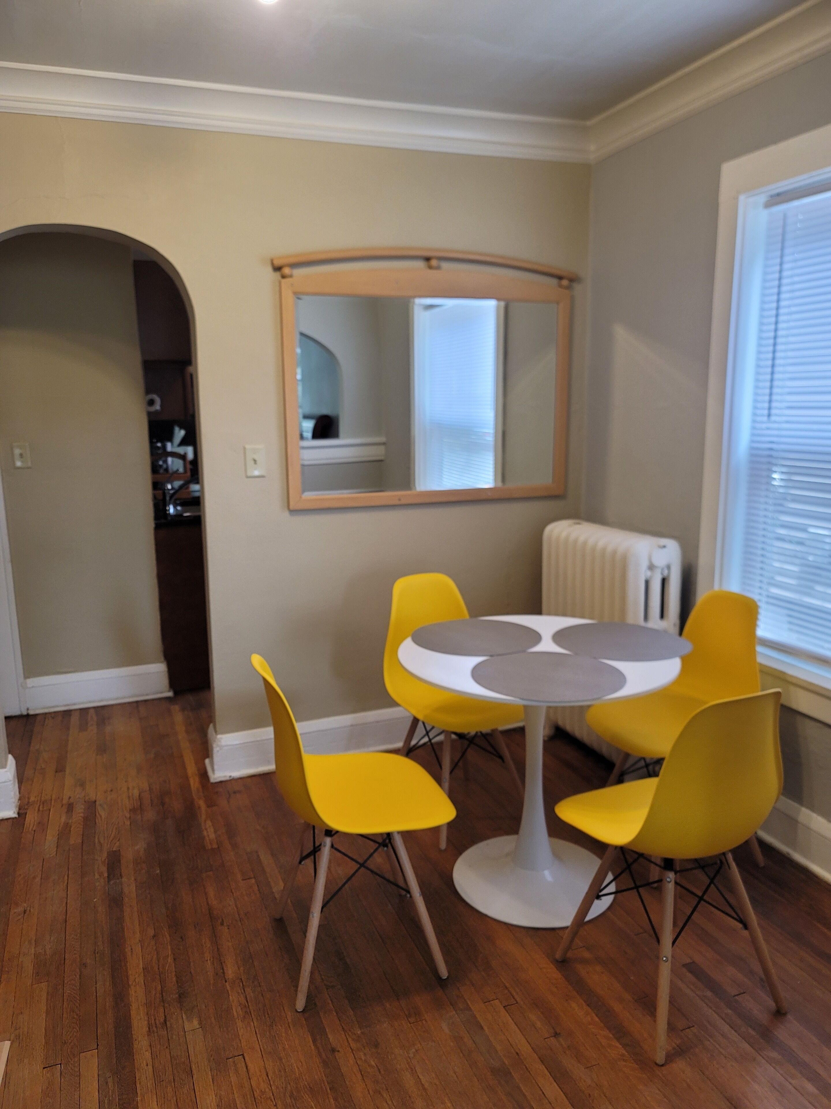 A dining area with a round table and four yellow chairs, a mirror on the wall, and a radiator.