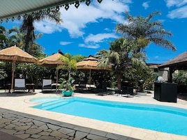 A swimming pool with lounge chairs and thatched umbrellas, surrounded by palm trees and a clear sky.