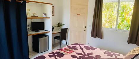 Quarto com cama, escrivaninha, cadeira, televisão e janela com cortinas.