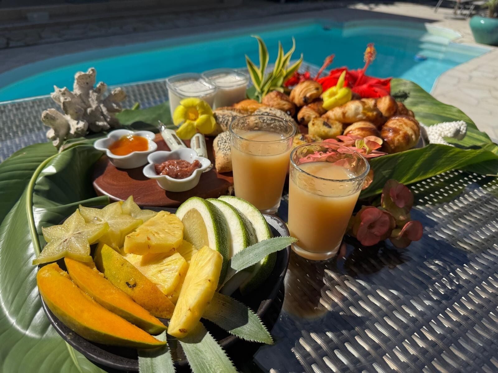 A tropical fruit platter with sliced mangoes and pineapple, accompanied by two glasses of a light-colored beverage, possibly a fruit juice or smoothie, and small bowls of condiments.