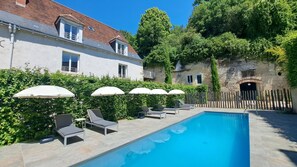 Ein Schwimmbecken mit Liegestühlen und Sonnenschirmen, umgeben von Grünpflanzen und einem Steinhaus.
